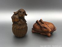 Photo of two Japanese netsuke carvings; a dog sitting on a basket and a reclining deer, both wooden with brown patina.
