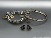Photo of two silver bracelets, one solid bangle with twisted rope detail and one charm chain bracelet, with pair of small floral clip-on earrings in front