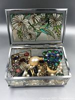 Full view of a glass jewelry box with assorted vintage jewelry inside including brooches, rings, and faux pearls.