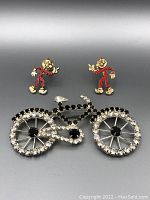 Photo showing two red and cream enamel cartoon character pins paired with a gemstone-adorned bicycle brooch on a neutral background.