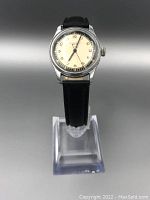 Front view of the vintage wristwatch on a clear stand showing cream dial, Arabic numerals, silver-tone bezel, and black leather strap.