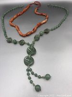Photo showing both necklaces laid out with the green jade necklace featuring various bead shapes and a pendant, alongside the orange carnelian beaded necklace.