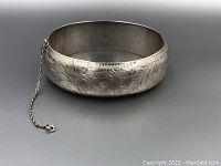 Closed view of a sterling silver bangle bracelet with floral engravings and a safety chain attached.