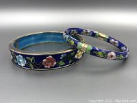 Photo showing two cloisonné bracelets with detailed floral enamel work on blue backgrounds, one wide and one narrow.