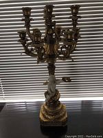 Decorative antique gold finish 10-light candelabra photographed against window blinds, showing full height and ornate detailing including sculptural elements at base and multiple candle holders.