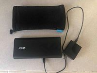 Photo of Anker branded portable battery pack connected to black wall charger power brick with cable, black protective pouch visible above.