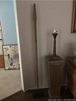 Full view of the antique wooden wagon tongue mounted vertically on a black base, positioned indoors next to a lamp.