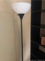 Full view of the torchiere lamp showing the white plastic shade atop a black pole against a beige wall.
