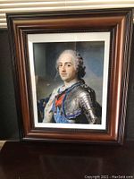 Framed print showing a portrait of Louis XV in armor within a dark brown wooden frame, photographed with natural lighting.