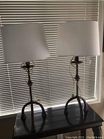 Two metal base table lamps with tripod-style legs and white drum shades, sitting on a dark surface in front of window blinds.