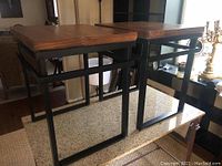 Pair of end tables shown side by side, highlighting their dark metal bases and wooden tops sitting on a granite countertop.