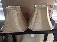 Pair of beige lamp shades on a table, showing front and side views to showcase shape and fabric texture.