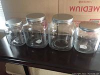 Four clear glass canisters with silver metal lids displayed on a dark table, two large and two smaller varying in size, all in good condition with no cracks or chips.