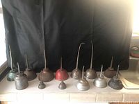 Thirteen vintage metal oil cans arranged in a row showing various shapes and conditions against a black backdrop. Includes different colors and sizes with visible rust and wear.