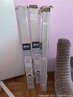 Three boxes of Bali Grab-n-go vinyl blinds standing upright against a pink wall, one with a blind partially exposed showing white slats and mounting hardware included.