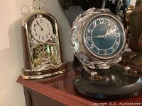 Two clocks on a wooden surface: vintage art deco style glass table clock with blue dial and a brass tone anniversary clock.
