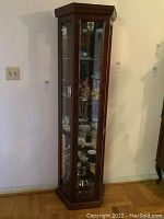Full front view of the tall, narrow wood curio cabinet with glass front and visible glass shelves and mirror back panel.