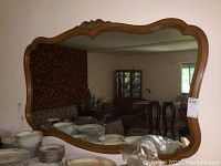 Photo of large wall mirror on wall reflecting room interior. Shows ornate wooden frame with carved, scalloped edges.