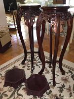 Two tall wood plant stands with carved floral aprons and curved legs, shown with two hexagonal wood bases on the floor nearby.