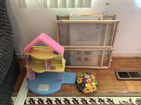 Wide shot showing Fisher Price playhouse, VTech toy, and stacked Supergate pet gates in background on wooden floor.