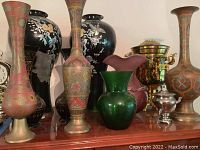 Cluster of brass vases, Asian style black urns with mother of pearl detailing, green and purple glass vases, and brass and stainless steel samovars arranged together on a wooden surface.