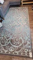 Full view of the rectangular blue and gray area rug with decorative floral and swirl patterns on wood floor near sofa and furniture.