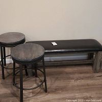 Rectangular entry way bench with black padded seat and two round wooden bar stools with black metal frames placed next to it on wood floor against wall.