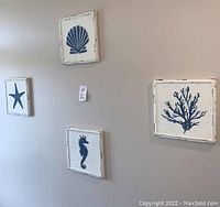 Four nautical themed square wall hangings with distressed white wood frames and navy blue designs (starfish, shell, seahorse, coral) on a beige wall.