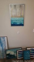 Photo of six various sized beach themed canvas paintings arranged against a wall, mostly showing blues and beige color tones, partly framed.
