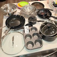 Wide view of all cooking ware items displayed on kitchen counter, showing frying pans, pots, mixing bowls, plastic plates, muffin tray, and strainers.