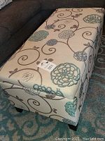 Full view of rectangular floral pattern storage ottoman showing beige fabric with teal and brown embroidered flowers and swirls, brown wooden legs visible.