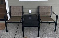 Front view of two black metal outdoor chairs with tan fabric seating and black glass top coffee table between them on a concrete patio surface.
