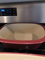 Large rectangular Pampered Chef ceramic casserole dish with reddish exterior and cream interior on stove.