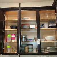 Kitchen cabinet with shelves holding plates, bowls, glass and plastic storage containers, various cups and shakers.