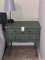 Side view of grey green side table showing three drawers and the black table lamp with grey shade on top, placed beside a bed on carpeted floor.