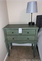 Grey-green side table with three closed drawers and table lamp with grey shade on top next to a bed.