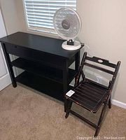 Picture showing the black wooden table with two shelves and pull-out drawers, white Sunbeam fan on top, and distressed black folding chair beside it.