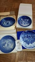 Four blue and white Bing & Grondahl collector plates, each depicting a different Christmas-themed scene and the year of release. Three plates are outside their boxes, one is shown in a protective open box.