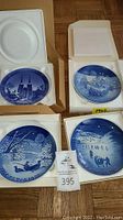 Four Bing & Grondahl collector plates shown in open Styrofoam packaging and cardboard boxes. Plates are blue and white porcelain with winter and Christmas scenes, some with visible dates 1967, 1970, 1972.