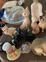 Assorted cat and kitten figurines including ceramic and glass cats, some black glass cats, and one gray sleeping cat on a glass base. Also includes a kitten playing with yarn and a figurine of a cat with a blue collar.