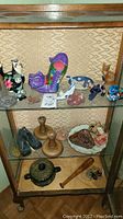 Glass display cabinet shelf lining with eclectic mix of colorful cat figurines, small boots, wooden candle holders, pottery dishes and small decorative items.