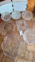 Multiple glass serving platters and bowls displayed on a wooden parquet floor including round platters with decorative patterns and a square platter with detailed designs.