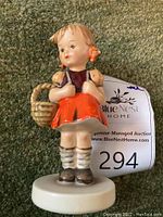 Front view of Hummel "School Girl" figurine standing on white round base, showing the girl holding a basket with a neutral expression and orange bows in hair.