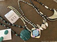 Four statement necklaces and one necklace with matching earrings displayed on a brown textured surface. The necklaces have large pendants featuring mother of pearl, wood, and resin in geometric patterns. The necklace and earring set is green carved stone.