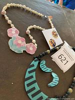 Photo of two necklaces, one with beige beads and a pink-blue floral pendant, and the other with black and beige beads and a turquoise geometric pendant, alongside pairs of matching earrings.