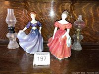 Two Royal Doulton porcelain figurines in purple and coral pink dresses, each about 8 inches tall, next to two small glass oil lamps on a wooden surface.