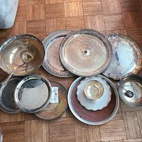 Multiple silver plated serving plates and platters arranged on floor showing variety of sizes, shapes, and decorative patterns including hammered and ornate designs.