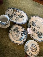 Five porcelain pieces laid on green carpet, showing platter, serving bowl, covered dish with lid, and small vase all with blue floral and gold-tone accents