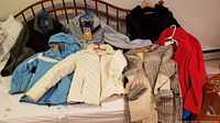 Full overview of the lot showing various jackets and coats laid out on a bed including blue Columbia windbreaker, cream quilted jacket, patterned wool sweater, and others.