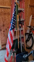 Photo showing American flag attached to green metal pole beside multiple garden rakes with wooden handles and a snow shovel.
